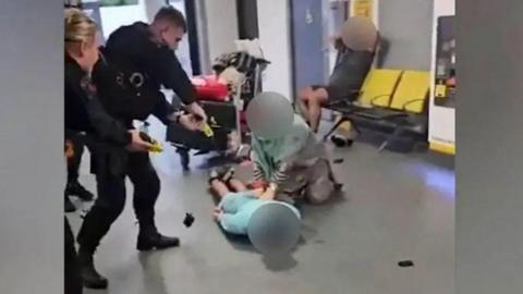 Two police officers point a laser at a man on the floor of an airport