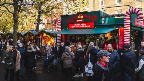 People walk past and queue at various stalls during York Christmas Markey, with attendees wrapping up in warm winter clothing. A mulled wine and hot chocolate stand is serving customers, with smaller craft stands nearby. 