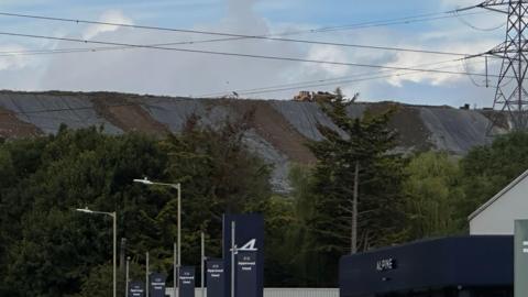 A pile of rubbish towers above some trees and lampposts. A truck sits on top on the distance