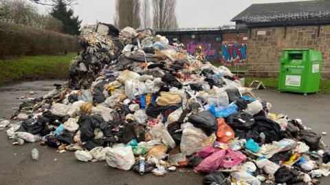 Pile of waste - which had caught fire in a bin lorry - in a car park. A green clothing bank is in the back of the picture towards the right, placed between the pile of rubbish and a stone building.