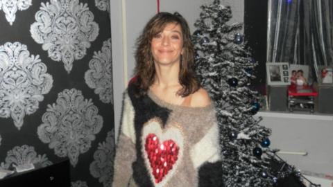 A woman with wavy shoulder length dark hair and an off the shoulder wooly jumper with a red love heart on the front stands smiling in front of a Christmas tree.