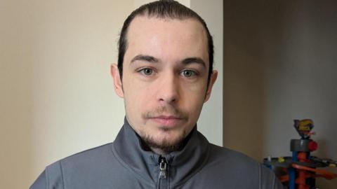 A young man with dark hair in a ponytail looking towards the camera.