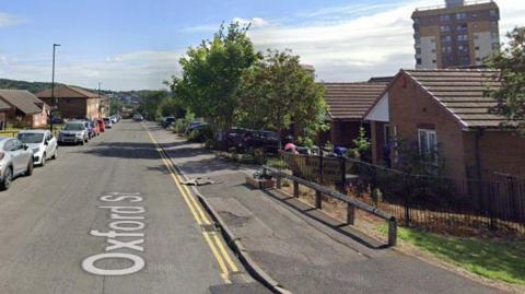 A street scene with bungalows on the right-hand side of the road and a row of cars parked on the left-hand side.