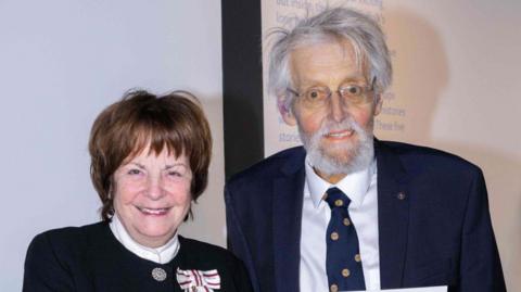 John Norman in a suit and tie with a woman beside him