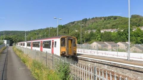 Abercynon train station. A white train with red doors is pulled up at the platform, with a metal fence on the other side of the tracks. A hillside covered in trees can be seen behind it. 