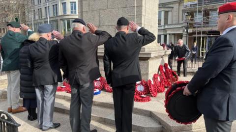 A general view of the Cenotaph in Jersey. Veterans are seen from behind. They are wearing suits and saluting. Wreaths are laid at the bottom of the cenotaph.