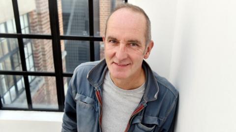 A short-haired man wearing a blue jacket and grey t-shirt, standing in front of a black-framed window with the side of an external wall visible outside. The room is white.