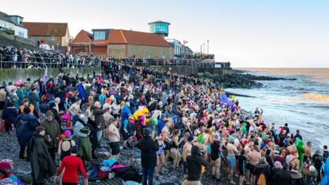 Hundreds of people on a beach. Some people are wearing their swimsuits while other people are wearing big winter coats. They are all facing the north sea. There are even more people onlooking by the pier.