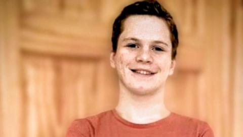 Joshua Robbins looks straight at the camera, whilst crossing his arms and smiling He's wearing an orange t shirt over a long black sleeved top. 