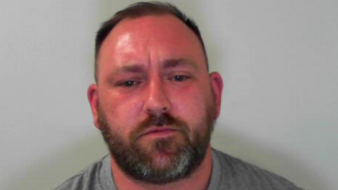 A custody photograph showing a man against a plain grey background. The man is facing the camera and has short, dark hair, closely cropped at the sides, with a beard and moustache. He is wearing a plain grey crew-neck T-shirt.