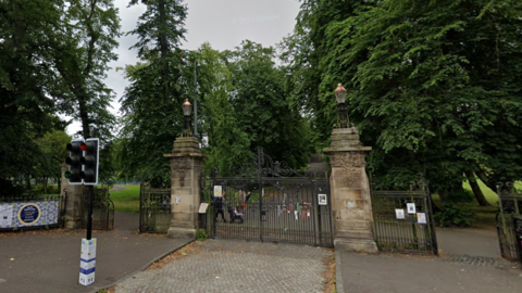The entrance to a park, with gates in front of a path and large trees