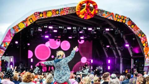Crowds gather in front of the Camp Bestival main stage which is lit with pink spotlights. The arched canopy over the stage has a CND logo at the top. A small child is sitting on someone's shoulders and waving their hands in the air.