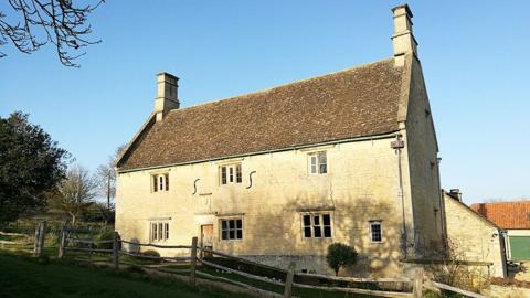 The outside of Woolsthorpe Manor, a 17th Century two-storey building of light stone and shingle tiles. It has two chimney stacks and a simple wooden bar fence and gate in front of it. The photograph is taken on a sunny day and trees are casting a shadow on the building.