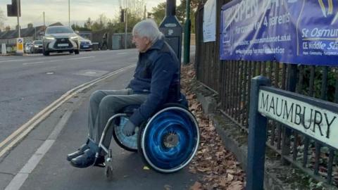 Geoff, a man with grey hair in a wheelchair with blue wheels. He is waiting on a pavement to cross a road. In the foreground is a road sign which reads: "Maumbury".