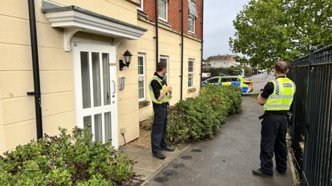 Two police officers wearing hi-vis yellow vests and black trousers and boots, standing outside the front door of a residential block of flats. There are bushes planted outside the windows and a metal fence. In the distance you can see a police car parked on the road.