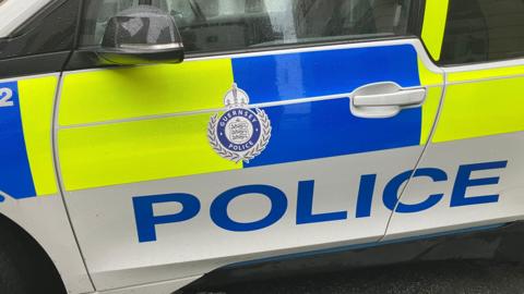 A close up shot of the door of a Guernsey Police car parked on a street. It is blue, white and yellow. It has police written in large blue block capitals on its side. Rain drops which have settled on the wing mirror, window and door are visible.