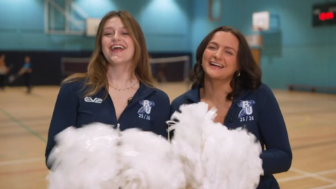Eve Kimberley (left) and Liv Teague are wearing dark blue cheerleaders outfits and waving pompoms in a gym.