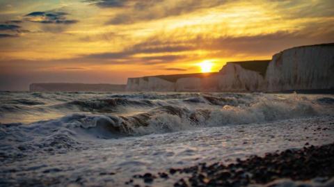 a close up of waves rolling into the shore with the sun shining in the background