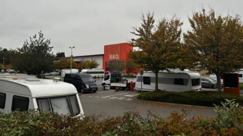 Several caravans parked in a car park outside a B&Q store in Patchway, near Bristol. There are trees and hedges in the car park and the orange storefront can be seen in the distance.