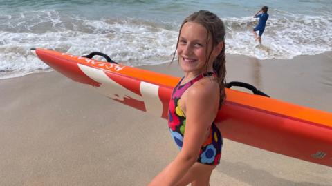 A young girl with a heavy orange surf board under her right arm prepares to go for a paddle