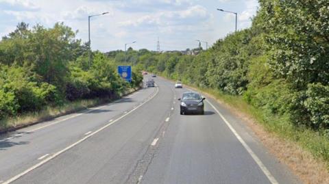 The dual carriageway shows several cars driving along the road on a sunny day. Hedges and trees line the road.