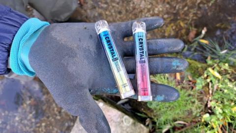 A rubber gloved hand holding two disposable vapes