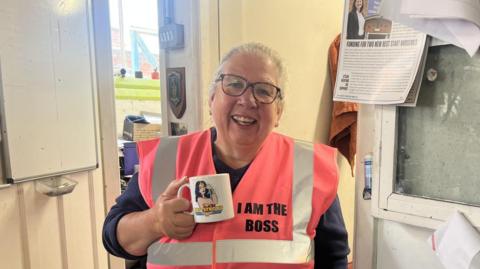 Karen Davies, a woman with pulled back grey hair, black rimmed glasses is smiling and holding a mug with a picture of Wonder Woman. Karen is wearing a peach flourescent vest with the words 'I am the boss' on her chest