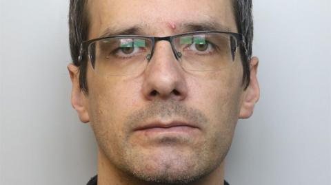 John Rigby has black hair that is turning grey. He is photographed wearing glasses.