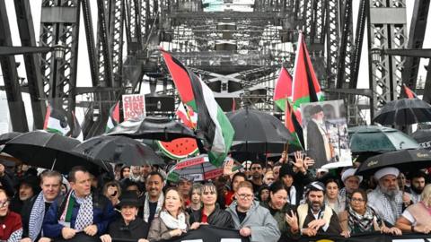 Hundreds of protesters gather under the arches of the Sydney Harbour Bridge, wearing raincoats and holding red, green, black and white flags. 