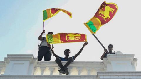 Three young men wave Sri Lankan fans from atop a white concrete balustrade