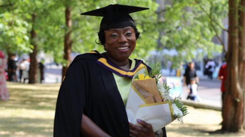 Makagbeh has dark hair and green eyeshadow. Smiling for the camera, she wears a green dress and a black mortar board cap and gown.