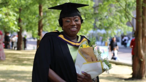 Makagbeh has dark hair and green eyeshadow. Smiling for the camera, she wears a green dress and a black mortar board cap and gown.