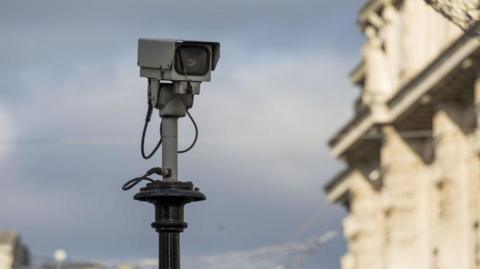 A stock image of a grey CCTV camera is on top a black pole. In the background there is blue sky and an out of focus building. 