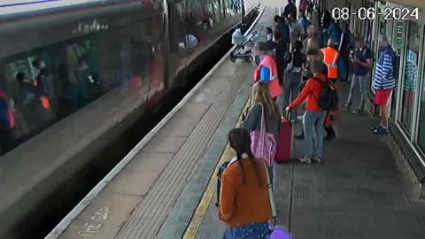 CCTV of a train arriving at a busy rail station platform with about 15 passengers all standing behind the yellow line but the pram pointing towards the train close to the platform edge.