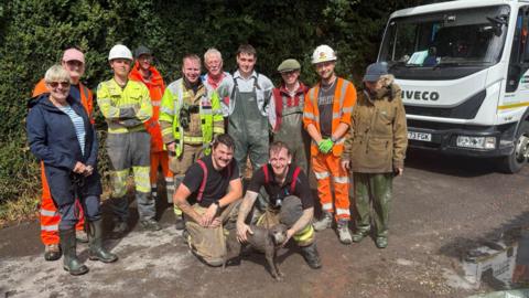 A group photo showing firefighters, council workers in hi-vis and local people in waders standing around a small dog who is covered in grey mud. They pose for a group photo on a gravel road and there is a white Iveco truck to their right
