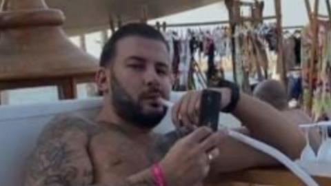 James Harding smoking a cigaratte and looking at his phone with his top off