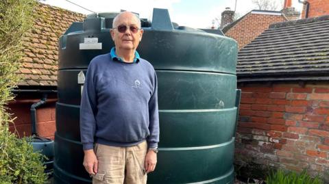 Chris Wheeler stands next to his dark green heating oil tank in his garden. He is wearing glasses, a blue sweatshirt and beige cargo trousers. The cylinder-shaped tank is in the corner of the garden in front of an old brick wall.