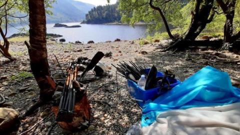 A collection of abandoned equipment on the shore of a large lake. Tent poles and pegs gathered together in boxes on the pebbled ground. Trees surround the equipment.