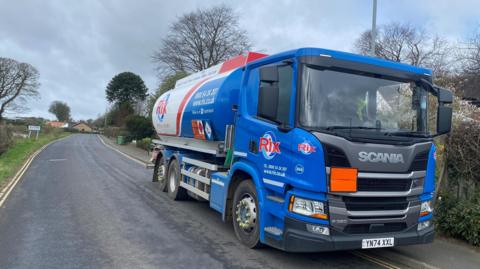 An oil tanker is parked on a rural road. It's a large blue and white lorry with Rix on the side. In the distance there is a sign saying Bilton and a couple of houses 
