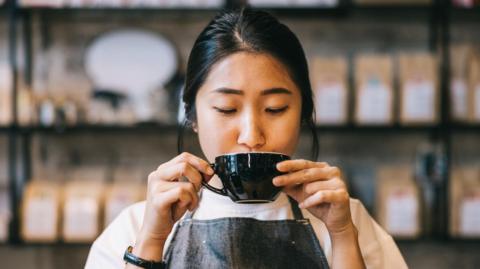 A woman in a dark grey apron sips on a cup of coffee.