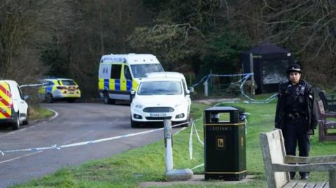 A number of police vehicles are at the scene of the incident, with a cordon in place and a police officer standing just outside the cordon. A number of benches can also be seen and a dark blue forensics tent is in place