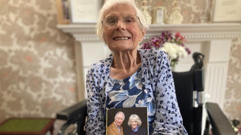 Elderly lady in a blue floral dress holding a card showing King Charles III and Queen Camilla. 