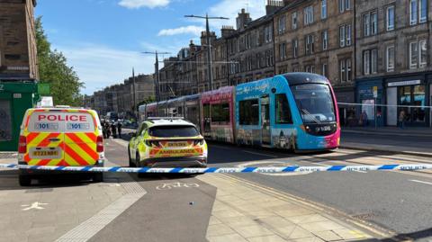A city street scene cordoned off by emergency services on a sunny day. Blue-and-white police tape stretches across the foreground, blocking the road. Behind the tape, a marked police van with high-visibility red and yellow chevrons and the word “POLICE” is parked on the left, next to an ambulance response car with yellow and green markings. A modern tram in bright blue and pink livery is stopped on tracks running through the middle of the street.
The street is lined with multi-storey stone buildings featuring rows of windows, chimneys, and ground-floor shops, including signs such as “The Leith Walk Gallery” and “Greet the Ch…” (partially obscured). Overhead tram wires and poles run along the street. A few pedestrians are visible on the pavement behind the cordon, and some people can be seen further up the road near the emergency vehicles.
The sky is clear blue with scattered clouds, and strong sunlight casts long shadows across the pavement and road surface. Yellow road markings and tactile paving are visible in the foreground near the blocked crossing. The scene suggests an active incident with the area restricted for safety.
