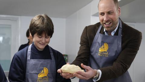 Prince William is holding a pasty. He is wearing a brown jacket and a blue apron.