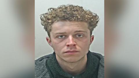 Police custody image of Frankie Bromley, who has light brown curly hair nd is wearing a black jumper and grey coat.