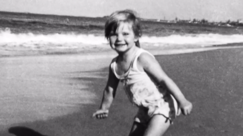 A black and white photograph showing three-year-old Cheryl Grimmer standing on a beach with waves gently lapping in the background. She has short fair hair and is wearing a white bathing costume. 