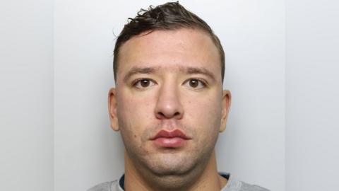 A custody image of a man with a round face, brown eyes and brown hair. He stares straight down the lens with a grey background behind him.