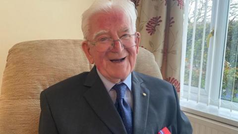 Bill Wake is smiling. He is a very elderly man wearing a suit with a collar and tie.