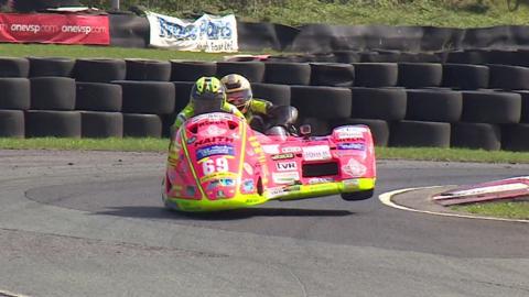 Nigel Limb in the side car of a pink motorcycle covered in sponsorship logos sits behind the rider as the bike comes round a corner with the sidecar wheel off the ground in front of a bank of stacked tyres. Both are wearing yellow racing leathers and helmets 