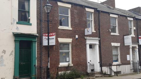 The outside of the terraced building which has a white door, one window on the ground floor and two on the first floor. It has a orange and blue logo on the windows and white sign above the door. The building on the left is cream with peeling paint. The building on the right has a white sign outside of it.